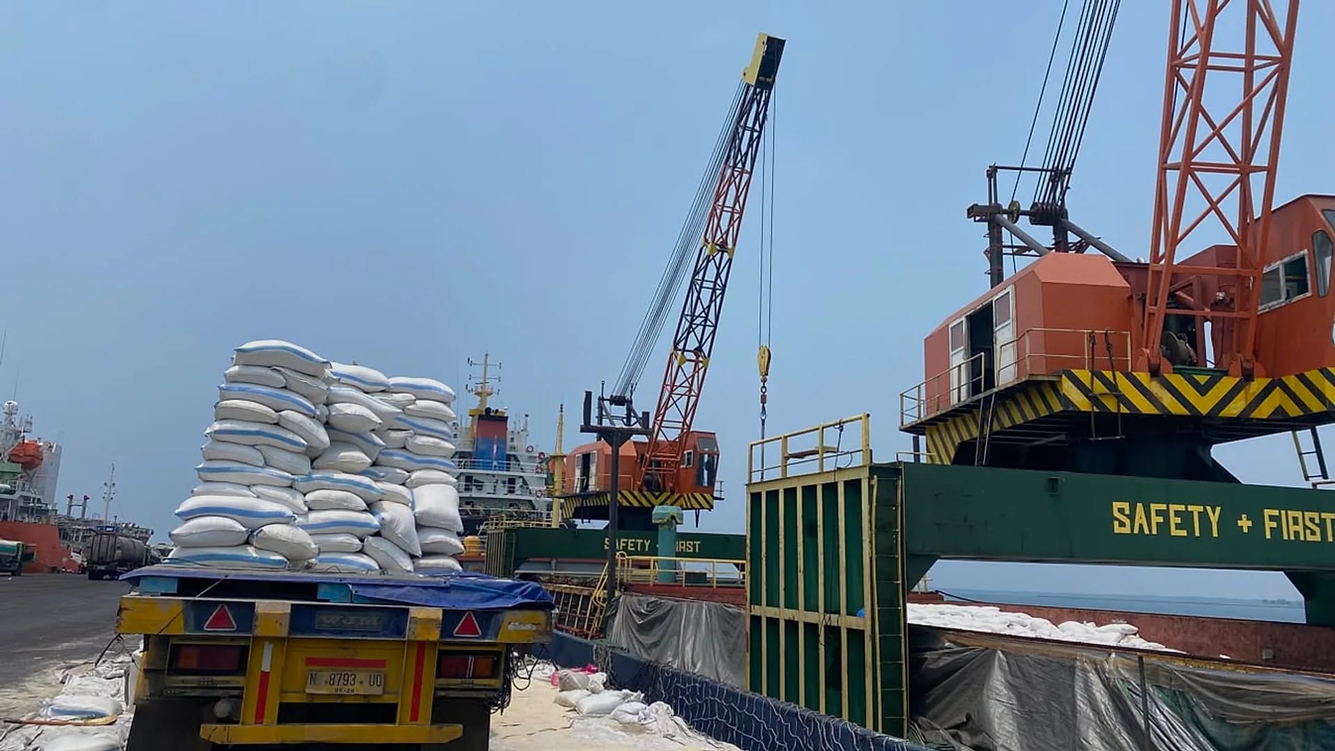Operasi bongkar muat di dermaga: truk bermuatan karung, crane pelabuhan, dan kapal kargo